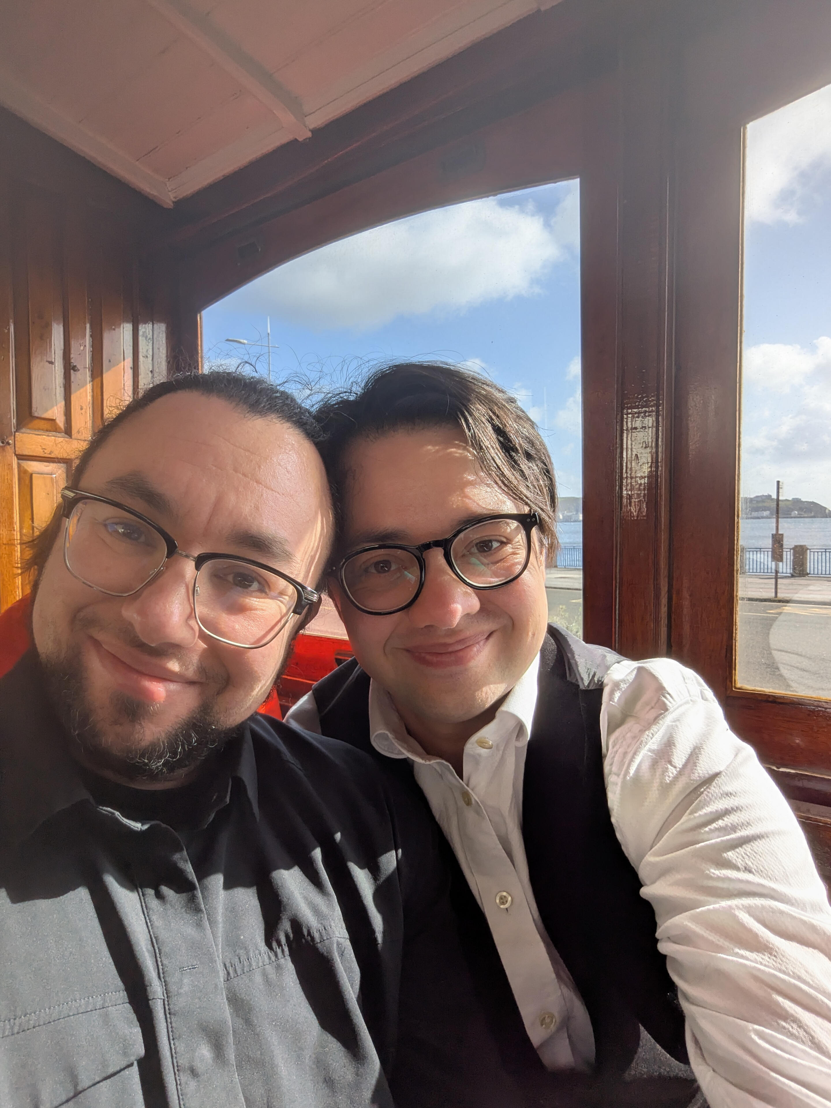 A selfie of Jack and Julian smiling while seated in an electric tram, the sea visible through the windows in the background.